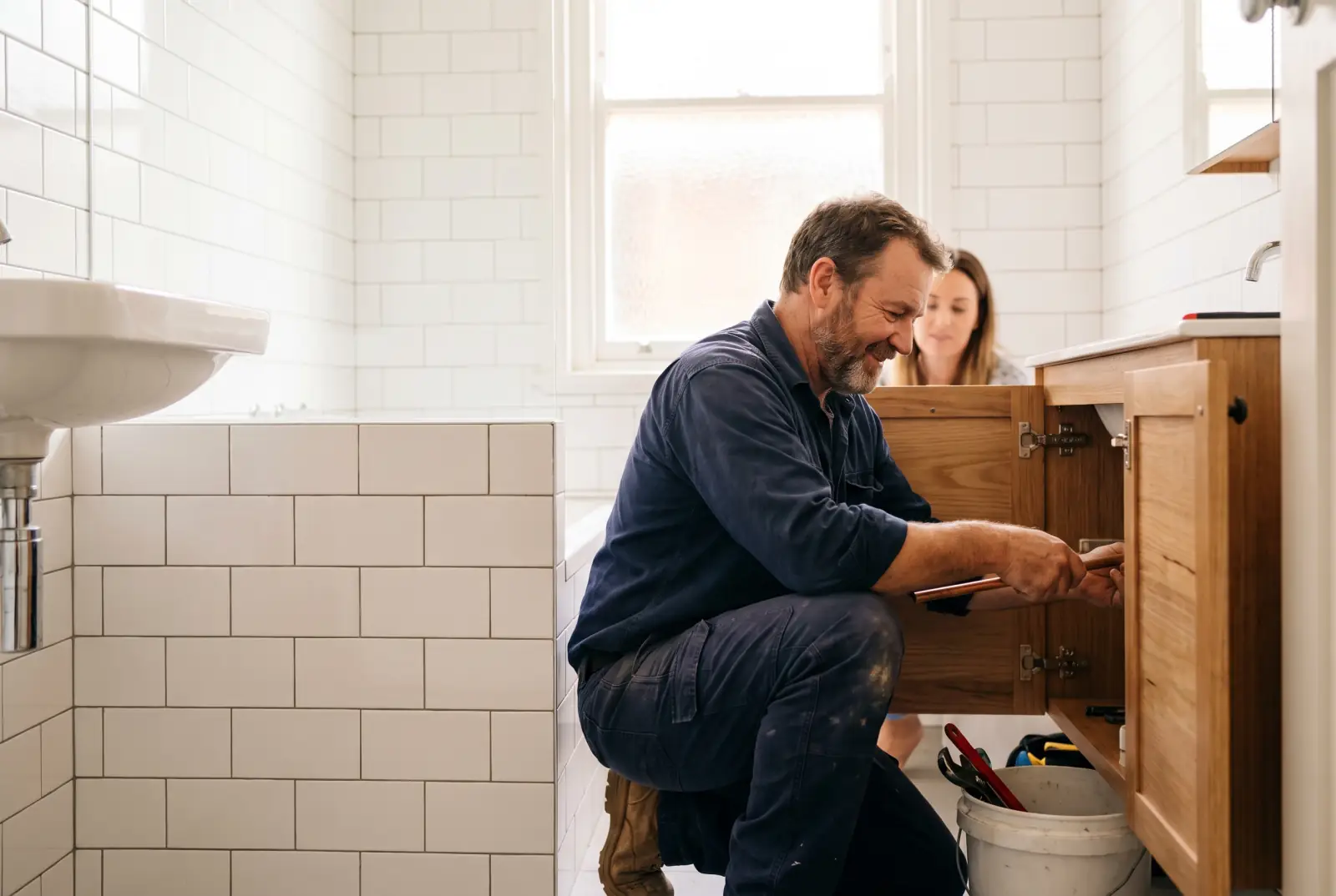 A plumber crouches to fit a pipe under a timber bathroom vanity in a renovated white-tiled bathroom